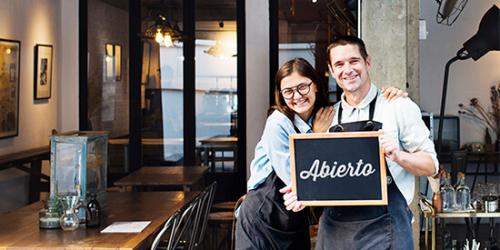 Promocion App ruralvia mi negocio - Dueños de una cafeteria sonriendo conel cartel de abierto mientras consultan la app ruralvia mi negocio en el movil Promocion App ruralvia mi negocio - Dueños de una cafeteria sonriendo conel cartel de abierto mientras consultan la app ruralvia mi negocio en el movil